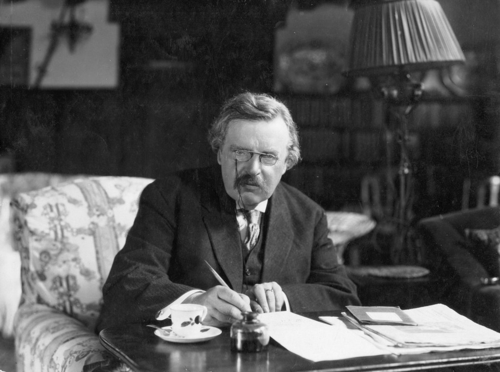 G K Chesterton writing at his desk.