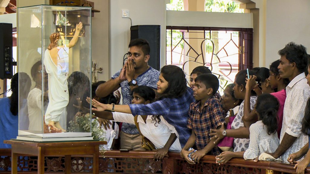 CAPTION (ACN-20190722-89394) Sri Lankans venerating and praying in front of the statue of Christ that is marked with the blood of the victims of the Easter Sunday attacks. It is now surrounded by glass and venerated in memory of those that lost their lives whilst worshipping on Easter Sunday 2019. Photo: Bartek Zytkowiak/Aid to the Church in Need.